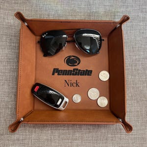 May include: A brown leather valet tray with a Penn State logo and the name Nick engraved on it. The tray has a pair of sunglasses, a car key, and several coins inside.