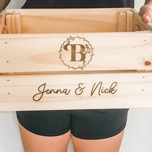 May include: A wooden crate with a personalized inscription. The crate is engraved with the letter "B" in a wreath and the words "Jenna & Nick".