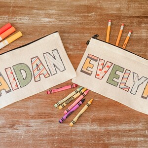 May include: Two white canvas pencil cases with zipper closures. The pencil case on the left has the name "AIDAN" written in colorful patterned letters. The pencil case on the right has the name "EVELYN" written in colorful patterned letters. There are several colored pencils and markers scattered around the pencil cases.