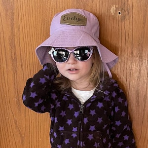 May include: A young girl wearing a purple bucket hat with a brown leather patch that says "Evelyn" on it. She is also wearing black and purple star patterned hoodie and pink sunglasses.