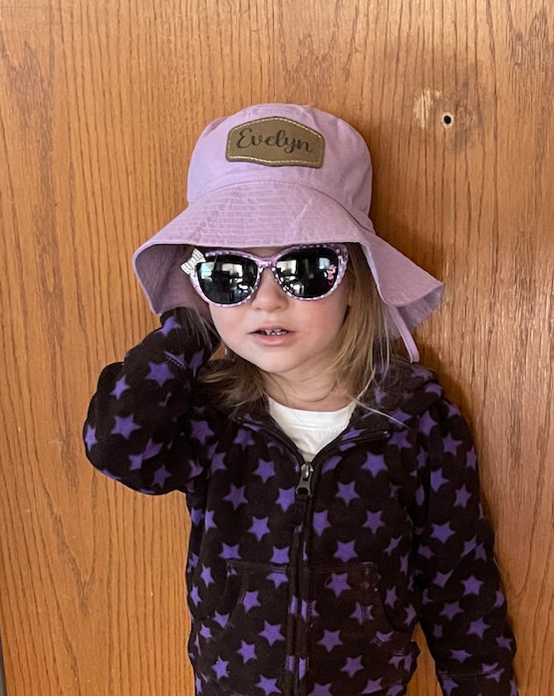 May include: A young girl wearing a purple bucket hat with a brown leather patch that says "Evelyn" on it. She is also wearing black and purple star patterned hoodie and pink sunglasses.