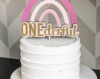 May include: A white cake with pink and white rainbow cake topper that says "ONEderful" in gold glitter letters.