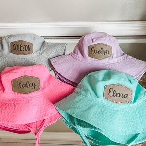 May include: Four personalized bucket hats in different colors. The hats are gray, purple, pink, and turquoise. Each hat has a brown leather patch with a name on it: Coleson, Evelyn, Haley, and Elena.