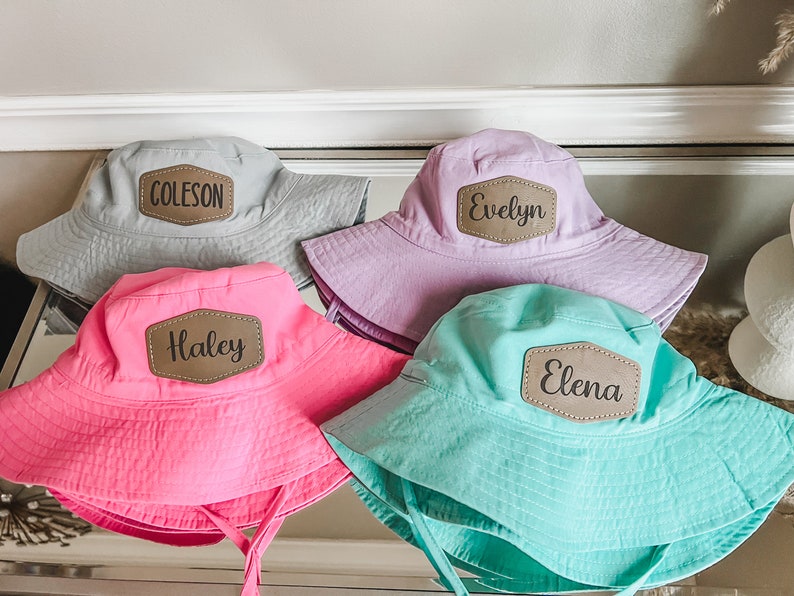 May include: Four personalized bucket hats in different colors. The hats are gray, purple, pink, and turquoise. Each hat has a brown leather patch with a name on it: Coleson, Evelyn, Haley, and Elena.