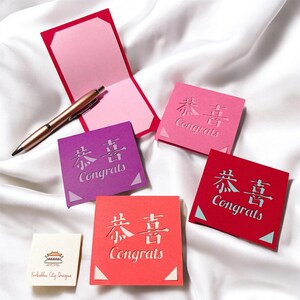 May include: Four square cards with the Chinese characters for "Congratulations" in a white cutout design. The cards are in pink, red, purple, and orange. The text "Congrats" is written below the Chinese characters on each card.