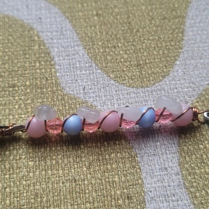May include: A silver hair clip with a beaded chain featuring pink, blue, and white beads. The chain is wrapped with copper wire and ends with a silver charm of a sun.