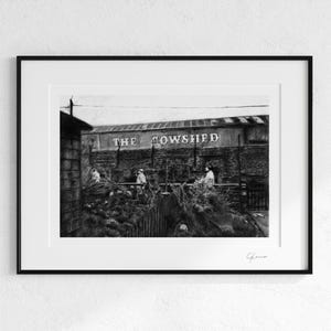 Könnte beinhalten: Schwarzweißfoto eines Gebäudes mit dem Schriftzug "THE COWSHED" an der Seite. Drei Personen stehen vor dem Gebäude.