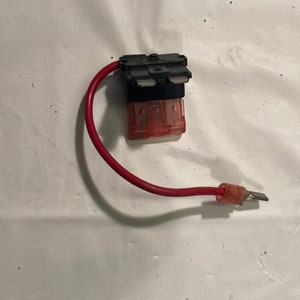 Può includere: Un filo rosso con un portafusibile nero e un fusibile rosso. Il portafusibile ha due contatti metallici e un corpo in plastica rossa.