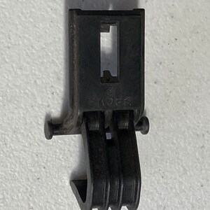 Puede incluir: Un clip de plástico negro con una sección superior rectangular y una sección inferior curvada y acanalada. El clip tiene una abertura central y dos pequeñas clavijas que sobresalen a cada lado. El material está marcado con el texto "POM 2".