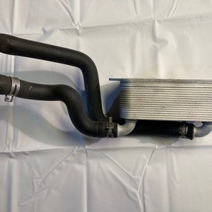 May include: An automotive oil cooler with black rubber hoses and metal clamps. The cooler has a rectangular, finned design and is made of silver-colored metal. The hoses are curved and connected to the cooler.
