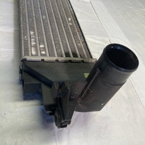May include: A silver radiator with a black plastic hose and a black plastic connector. The radiator has a series of fins and is designed to cool a vehicle's engine.