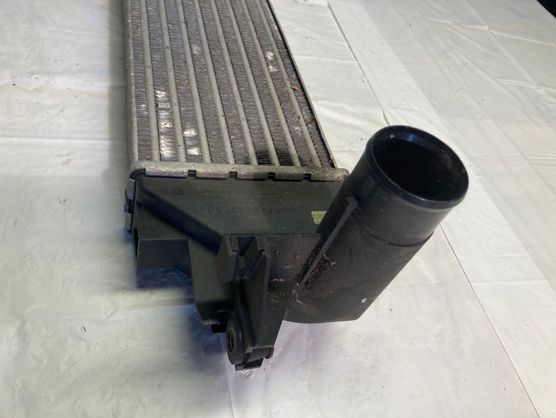 May include: A silver radiator with a black plastic hose and a black plastic connector. The radiator has a series of fins and is designed to cool a vehicle's engine.