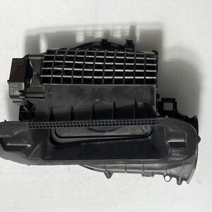 Puede incluir: Un componente de plástico negro para el aire acondicionado automotriz. La pieza tiene forma rectangular con una serie de ranuras paralelas en la parte superior. Una estructura negra en forma de correa recorre el lateral del componente.