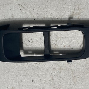 Puede incluir: Pieza de coche de plástico negro con dos aberturas rectangulares. La pieza está diseñada para contener interruptores o botones.