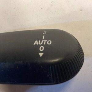 May include: Black plastic gear shifter with white lettering that reads "AUTO 0 1 2" with a white arrow pointing down.