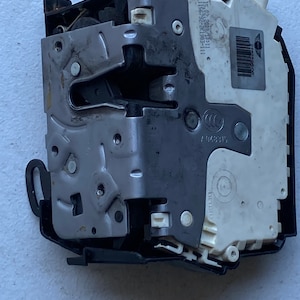 May include: A grey and black car door lock mechanism with a white plastic cover. The cover has a barcode and the text "51 02 - 4008 101 - 11 11 51 52 - 2K1 985 - 11" printed on it.
