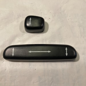 Op de afbeelding: Twee zwarte plastic knoppen met pijlen die de richtingen omhoog en omlaag aangeven. Deze knoppen worden gebruikt om de positie van een autostoel aan te passen.
