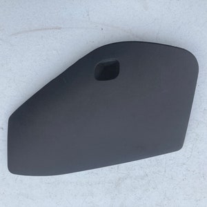 Puede incluir: Una pieza de coche de plástico negro con una pequeña abertura rectangular. La pieza es curva y tiene una superficie lisa.
