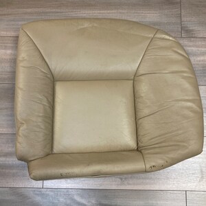 May include: A beige leather car seat with a worn, faded appearance. The seat has a rectangular shape with a raised center section and a slightly curved back.