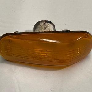 Puede incluir: Una luz de señal de giro de coche de plástico amarillo con una lente acanalada y una base de plástico blanco.