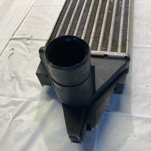 May include: Close-up of a black intercooler with a large, round, black inlet pipe. The intercooler has a silver, finned core.