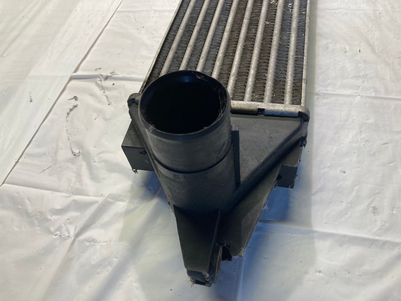 May include: Close-up of a black intercooler with a large, round, black inlet pipe. The intercooler has a silver, finned core.