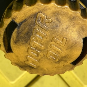 Peut inclure: Gros plan sur un bouchon en plastique jaune avec les mots "MOTOR OIL" en relief. Le bouchon est sale et présente des traces noires.