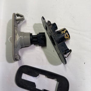 Puede incluir: Una pieza de automóvil desmontada con una carcasa de plástico gris, un conector de plástico negro y una base de plástico negro. La pieza tiene una forma rectangular y está diseñada para encajar en la carrocería de un automóvil.