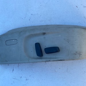 Puede incluir: Panel de control de asiento de coche de plástico beige con dos botones negros. El panel tiene una abertura rectangular con un botón rectangular más pequeño en el interior.