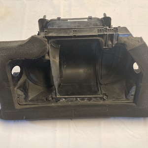 Puede incluir: Pieza de coche de plástico negro con aislamiento de espuma. La pieza tiene una forma rectangular con una gran abertura en el centro. La abertura está rodeada por un marco de plástico negro.