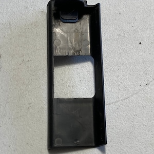 Puede incluir: Pieza de moldura de plástico negro con una abertura rectangular y varios agujeros circulares pequeños. La pieza de moldura tiene una pequeña pieza de plástico negro rectangular unida a la parte superior.