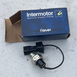 Peut inclure: Un capteur électrique noir avec un corps en métal argenté et un fil noir. Le capteur porte l'inscription "30. 7H". Le capteur est emballé dans une boîte bleue avec le texte "Intermotor Genuine Import Parts" et le logo "SMP".