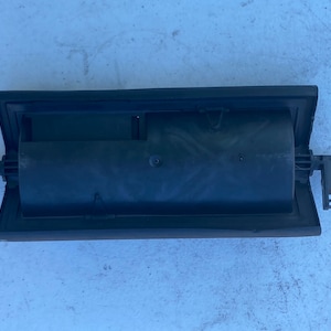 Puede incluir: Ventilación de aire de coche de plástico negro con una abertura rectangular y un panel deslizante. La ventilación tiene dos pequeñas pestañas a cada lado.