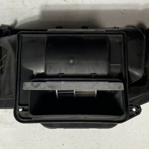 Puede incluir: Un conjunto de ventilación de aire automotriz de plástico negro. El conjunto tiene una forma rectangular con un área de ventilación abierta y una serie de componentes internos. La ventilación está diseñada para dirigir el flujo de aire.