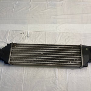 May include: A black metal intercooler with a silver radiator core. The intercooler has two black plastic end caps with inlets and outlets.