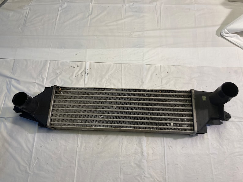 May include: A black metal intercooler with a silver radiator core. The intercooler has two black plastic end caps with inlets and outlets.