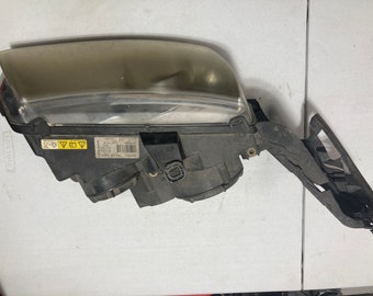 May include: A black and silver car headlight with a clear lens and a sticker that reads "MADE IN SLOVAKIA" and "DOT SAE", along with a warning symbol.