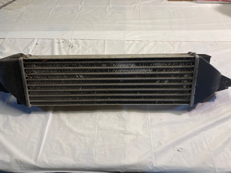May include: A silver intercooler with black end caps. The intercooler has a rectangular shape and is made of metal with a grid pattern.