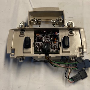 May include: A beige automotive interior light with a circuit board and two black buttons. The light has two wires connected to it, and the wires are connected to two black connectors.