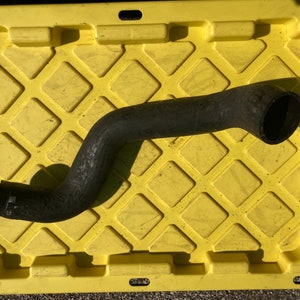 May include: A black, curved rubber hose, possibly for a vehicle, lies on a yellow plastic container. The hose has a textured surface and circular openings. The container has a diamond pattern.