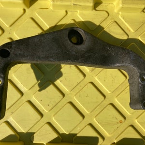 Könnte beinhalten: Eine gebrauchte, graue Metallhalterung mit gebogenem Design und mehreren kreisförmigen Löchern. Die Halterung hat einen U-förmigen Abschnitt und einen zentralen Befestigungspunkt. Wahrscheinlich ein Automobil- oder Industrieteil.