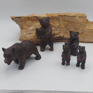 Op de afbeelding: Een verzameling handgesneden houten berenbeeldjes in verschillende poses. De beren zijn donkerbruin met zichtbare houtnerf en variëren in grootte van kleine welpen tot een grotere volwassen beer. Een stuk lichtbruin hout dient als achtergrond.