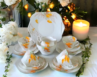 May include: A vintage tea set featuring white porcelain cups, saucers, and a serving plate, adorned with a delicate orange and beige floral pattern. The set includes teacups, saucers, a creamer, and a serving plate, all with gold trim. A candle and flowers are in the background.