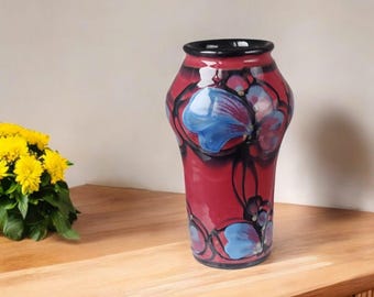 May include: A decorative vase with a red base and a black rim. The vase features a floral design with blue and purple accents. The vase is on a wooden surface.