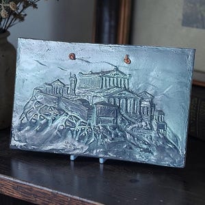 Puede incluir: Una escultura de relieve metálico rectangular que representa un complejo de edificios clásicos sobre una colina rocosa. La escultura es de color verde azulado apagado y tiene dos pequeños lazos para colgar. La pieza se exhibe sobre una superficie de madera oscura.