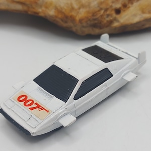 Könnte beinhalten: Ein weißes Spielzeugauto mit schwarzem Dach und Fenstern, mit der roten Nummer "007". Das Auto hat kleine Flügel an den Seiten und ist wahrscheinlich ein Sammlerstück.