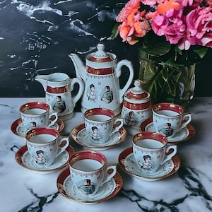 May include: A vintage porcelain tea set with a white and red design, featuring portraits and gold trim. The set includes a teapot, creamer, sugar bowl, and several teacups with saucers. A bouquet of pink flowers is in the background.