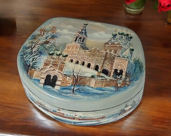 May include: A hand-painted, oval-shaped box with a winter scene depicting a church with multiple domes and towers. The box features a light blue background with snow and trees. The artwork is detailed and colorful.