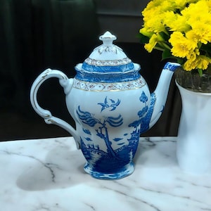 Op de afbeelding: Een wit en blauw porseleinen theepot met deksel, handvat en tuit. De theepot heeft een blauw landschapsontwerp met vogels, bomen en gebouwen. Het handvat en de tuit hebben gouden accenten.