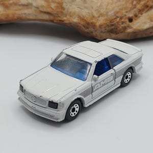 May include: A white toy car with black wheels and blue interior. The miniature vehicle has a sunroof and a detailed front grille. The car is a vintage model, showing signs of wear.